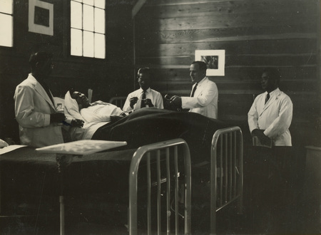 A black and white photo of medical personnel tending to a patient in a room. Four medical personnel surround a man on a bed.