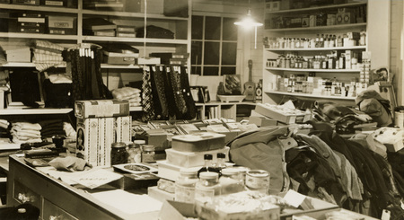 A black and white photograph of the merchandise inside of a store such as bottles, clothing, and food.
