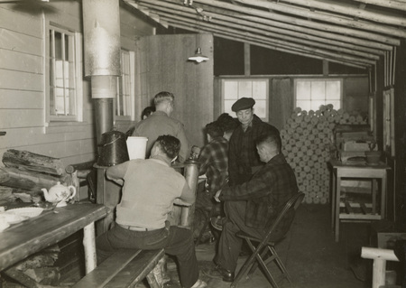 A black and white photograph of a group of men sitting around a wood-burning stove.