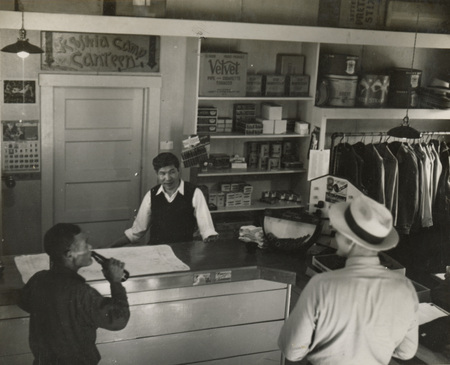 A black and white photograph of three men inside of a store with a sign reading "Kooskia Camp Canteen" on the wall.