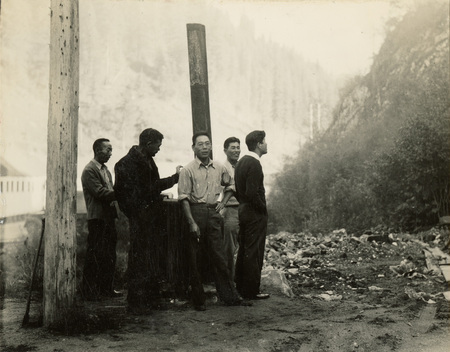 A black and white photograph of men standing outside next to two large posts. One of the men is smoking a cigarette and the other four are standing.