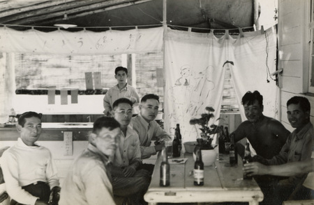 A black and white photograph of seven men sitting at a table on a covered patio. On the table are some bottles.