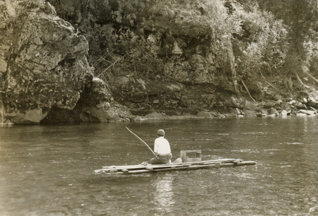 A black and white photograph of a man fishing on a raft in a river.