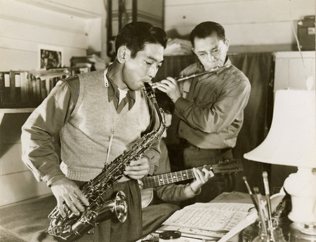 A black and white photograph of three men playing instruments: one playing a saxophone, one playing a guitar, and one playing a flute. 