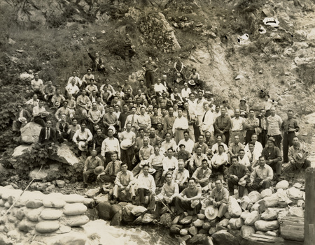 A group photograph of men at a worksite. There are many sandbags in front of the men who are sitting on the hill side.