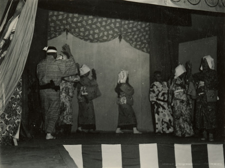 A black and white photograph of a performance on a stage. The men are dressed in traditional Japanese clothing.