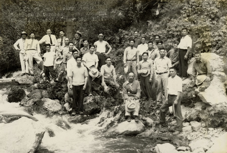 A black and white image of a large group of men next to a creek holding a sign that reads: "Anniversary Japanese Internees, Camp Kooskia, Kooskia Idaho, May 28, 1944". 