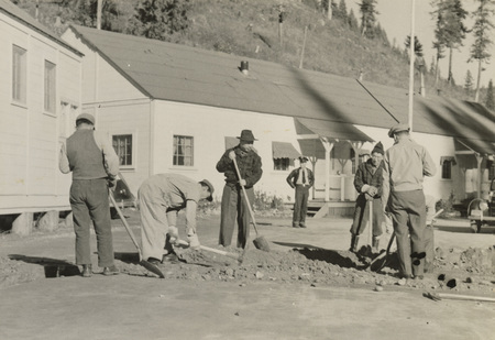 A black and white photograph of five men digging with shovels and pickaxes in front of buildings.