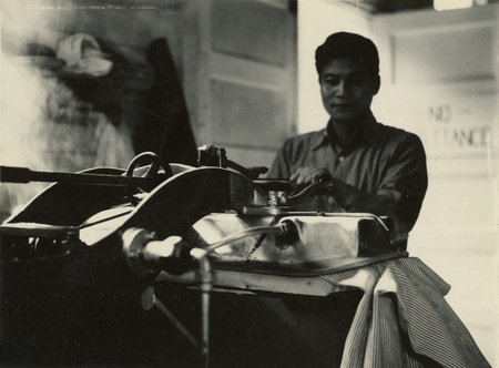 A black and white photograph of a man using a clothing press. The cloth in the press has a narrow striped pattern.