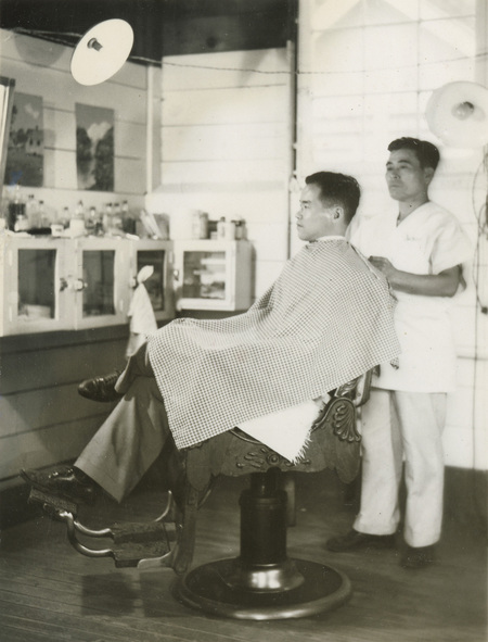 A black and white photograph of a man wearing a cape in a barber's chair. A male barber in all white stands behind the chair, to the right.