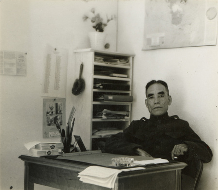 A black and white photograph of a man sitting at a desk. There is a bookshelf behind him and a map above him on the wall.