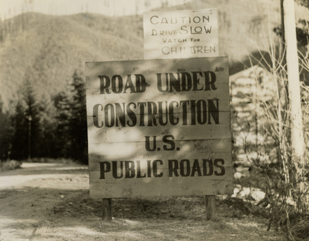 A black and white photograph of a sign reading "Caution: Drive SLOW, watch for children" and "Road Under Construction U.S. Public Roads".