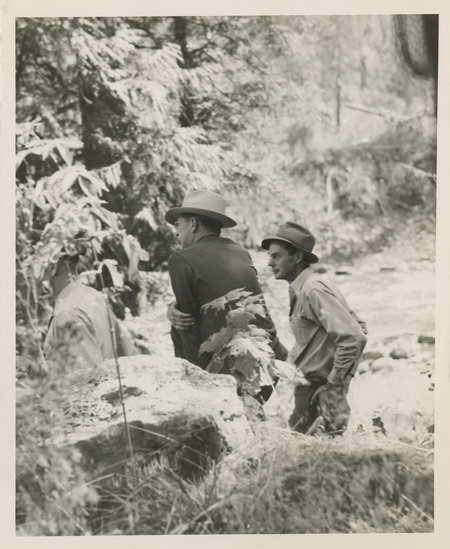 A black and white image of three men standing by a river, talking to each other. There is a large rock in front.