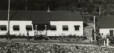 A black and white photograph of men walking in front of a long building.