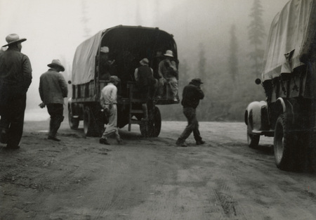 A black and white photograph of men standing behind a large transport truck. Men are also sitting inside of the truck.