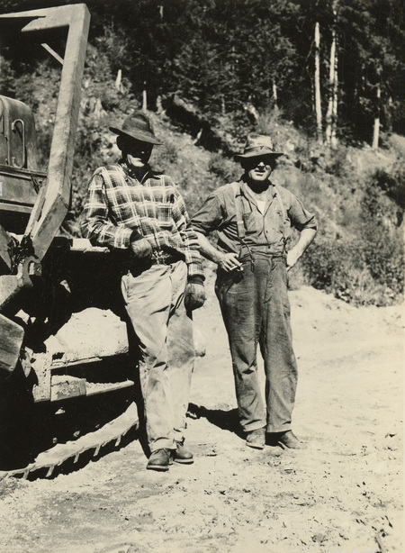 A black and white photograph of two men standing next to a bulldozer on a dirt road.