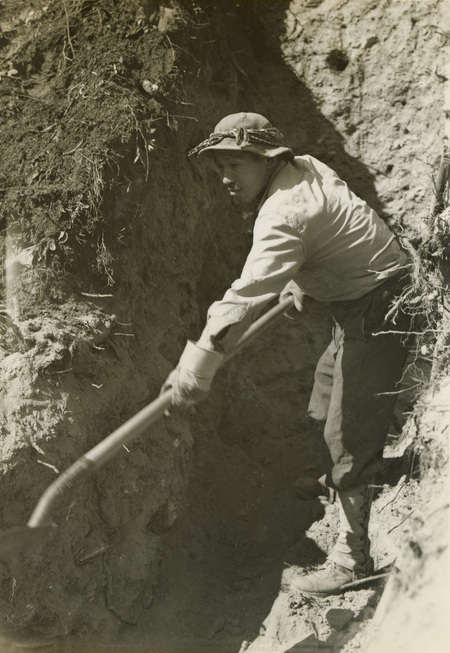 A black and white photograph of a man in a trench with a shovel.