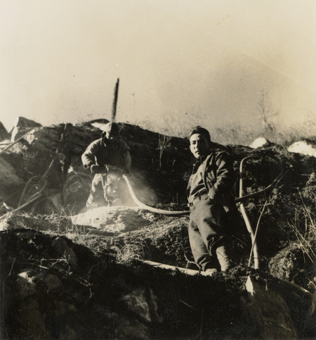 A black and white photogrpah of two men working on a hillside with construction tools, including a jackhammer.