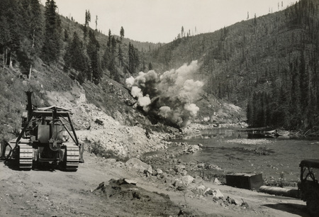 A black and white photograph of an explosion along a river, with construction images along the road where the explosion is occurring.