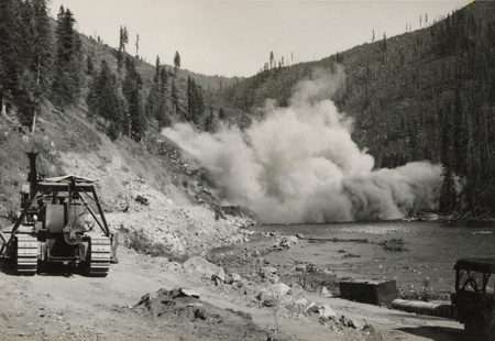 A black and white photograph of an explosion along a river, with construction images along the road where the explosion is occurring.