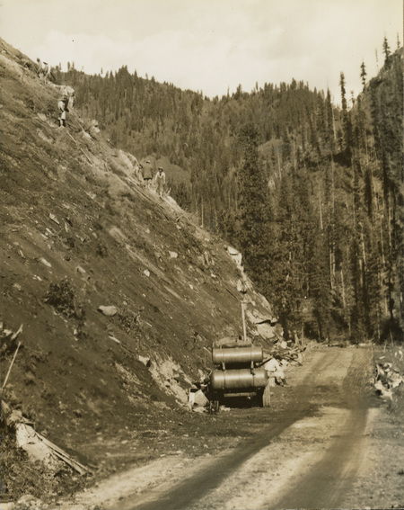 A black and white photograph of a hillside. Men are working on the hillside and construction equipment is parked on the dirt road below.