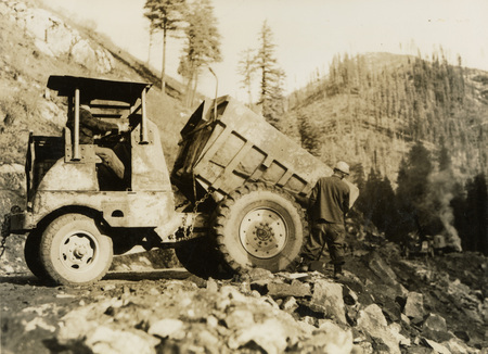 A black and white photograph of two men working with a dump truck on the edge of a hillside.