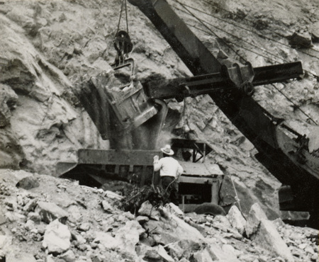 A black and white photograph of a crane moving dirt on a hillside. A man in a white shirt stands in front of the crane.