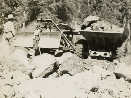 A black and white photograph of two men moving large rocks and boulders with construction equipment. There are two large truck-like pieces of equipment.