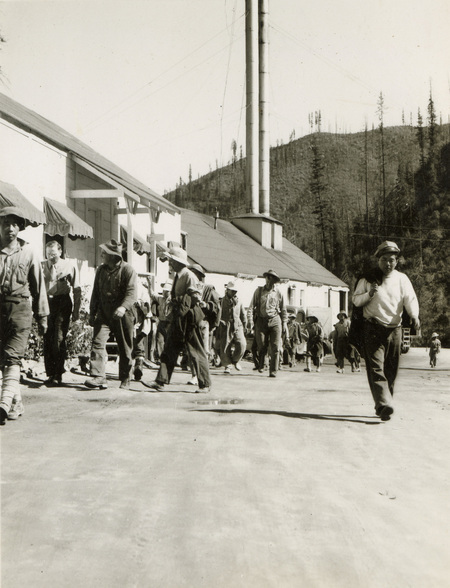 A black and white photograph of many men walking while looking at the side of a building. Most of the men are wearing helmets or hats.