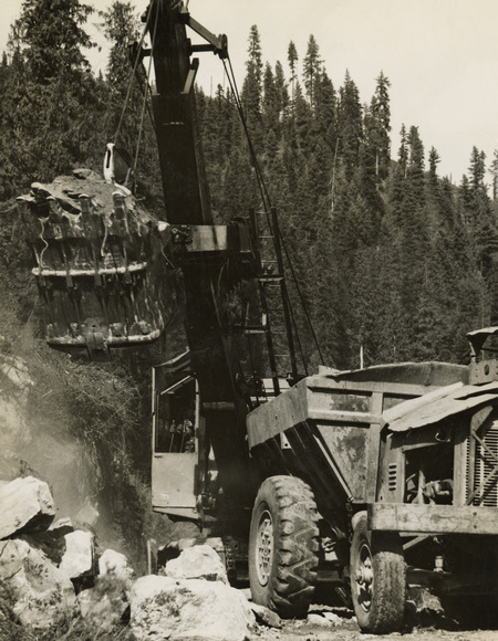 A black and white photograph of a crane and other construction equipment moving large rocks and dirt from the road.