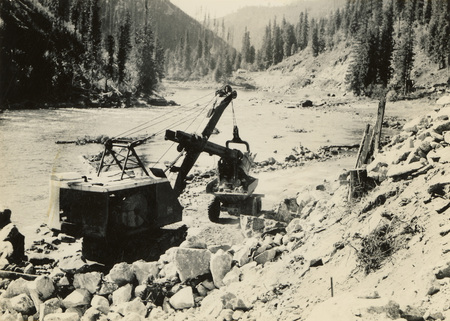 A black and white photograph of a crane with a large log in its grasp. The crane is next to a scenic view of a river.