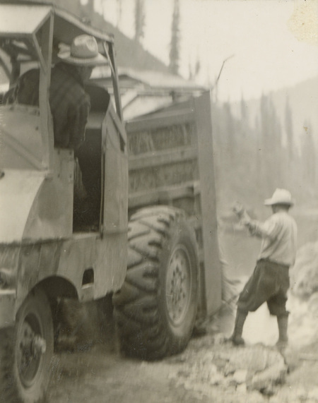 A black and white photograph of a dump truck. A man stands toward the back of the truck and is directing a man in the driver's seat of the truck.