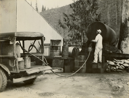A black and white photograph of a man outside filling a fuel tank and vehicle with fuel from a larger stationary tank.
