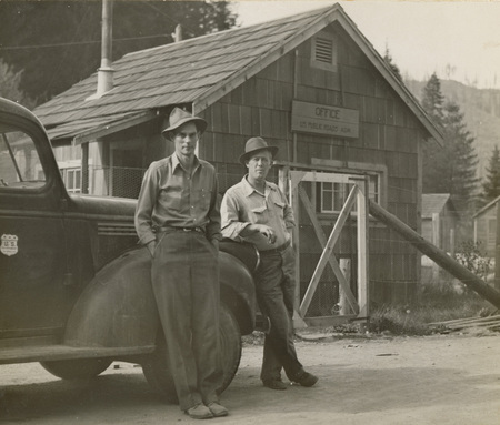 A black and white photograph of two men leaning against a vintage truck in front of a wood building.