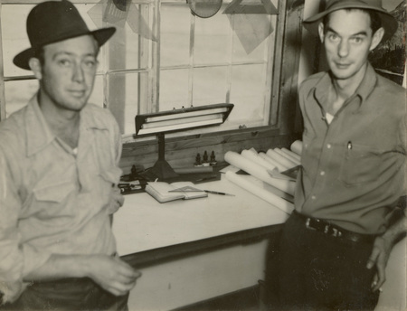 A black and white photograph of two men standing in front of a desk. There is a large lamp at the center of the desk and rolls of paper behind the man on the right.