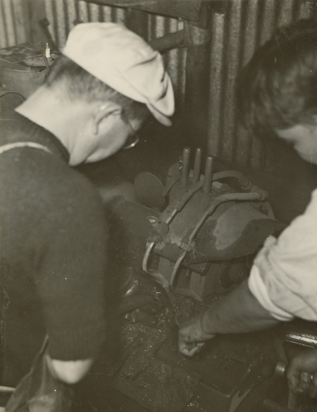 A black and white photograph of two men working on an engine in a machine shop. Their backs are to the camera.