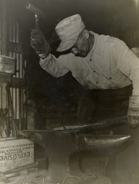 A black and white photograph of a man who is about to hit an anvil with a hammer.
