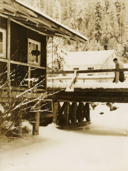 A black and white photograph of a man crossing a snow-covered bridge to a log building.