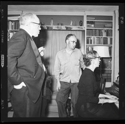 Woman plays the piano while people stand around her singing at a costume party.