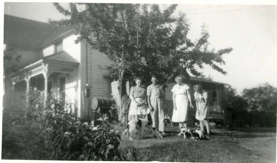 The Packard family in front of their farmhouses; the house is no longer standing near the meat locker. Individuals named from left to right: Julia Packard (from WIsconsin), Lyle, Irma Packard (from Wisconsin), Wilma Packard, and Tippy the dog and her pups.