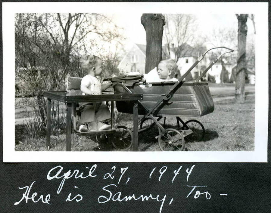 Photograph of Bill Hopkins, seated at table, and Sammy, in the pram, playing together outside.