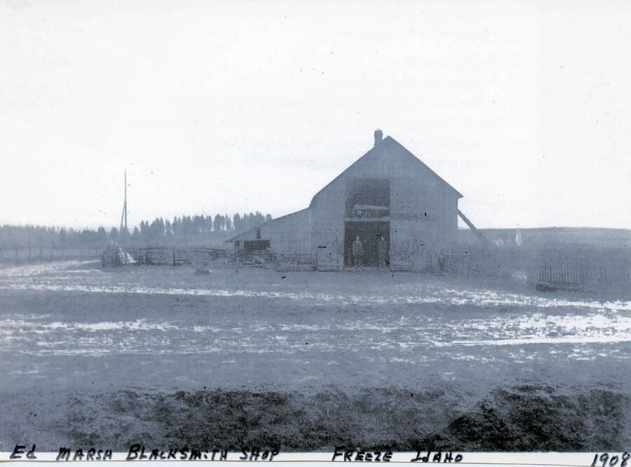 View of Ed Marsh's blacksmith shop in Freeze, Idaho. Two men can be seen standing in the doorway of the shop.