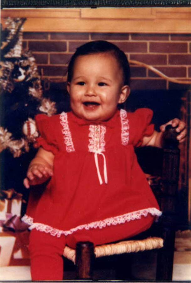 Shannen Fleener's 2nd Christmas. Shannen wears a red dress and sits on a chair in front of a fireplace.