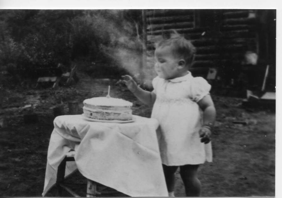 Unknown child's first birthday. The toddler stands beside a table on which a birthday cake with a single candle sits.