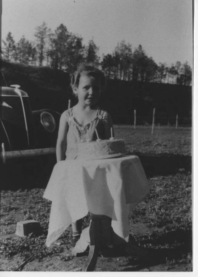 Joyce Fleener standing behind a table with her 3rd birthday cake sitting on top. A car can be seen parked behind her.