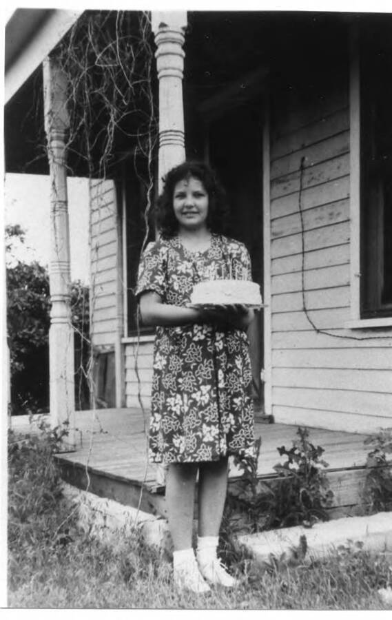Joyce Briney poses holding a cake for an 11th birthday photo in Beloit, Kansas in front of her grandmother, Grace Briney's, home. The photo was taken by Joyce's aunt, Melva Briney. Melva also made the banana cake, which was made with fresh bananas and whipped cream icing. This was the year that Joyce and her sister, Nyla June Briney, traveled to Kansas all by themselves via a Greyhound bus.