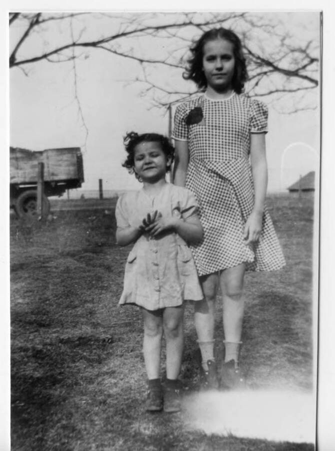 Della Mae and Linda Rosenfelt standing next to each other. The two girls are Briney family cousins. A truck and a field can be seen in the background behind the girls.