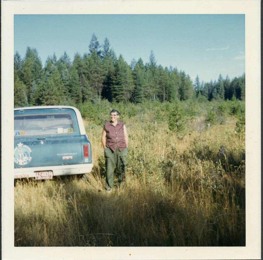 Orelia Kelly beside a blue pickup truck parked in the field. Tall pine trees are visible in the background.