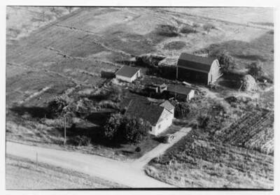 View of Vernon and Alice Briney homestead. The cleared fields that are in the background formerly contained cherry orchards. The Briney family would sell cherries to a canning facility in Post Falls, ID. They would take the back seat out of the car in the driveway to load cherry lugs, boxes approximately the size of what sweet cherries come in today, to take to the canning facility. Buildings, cars, and a haystack can be seen sitting on the lot.