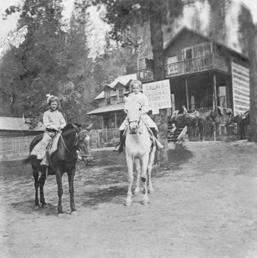Two young girls riding horses outside a group of houses.